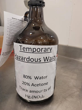 Improperly labeled day jar, does not have a depiction of the hazards and contains chemical abbreviations.