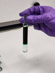 Test tube depicting a clear, unknown liquid that has sunk to the bottom of deionized water that was dyed green.