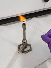 Picture depicting a cotton swab with unknown solid material in a Bunsen burner flame.  Cotton swab is emitting orange and yellow flames, indicating a positive flammability test.
