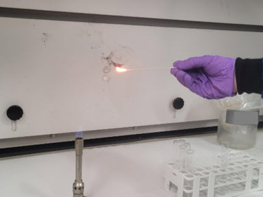 Picture depicting lab personnel in purple nitrile gloves holding a cotton swab over a Bunsen burner. The swab is on fire and emitting black smoke, indicating a positive flammability test.